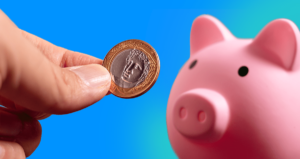 Mão segurando moeda de um real em frente a um porquinho
