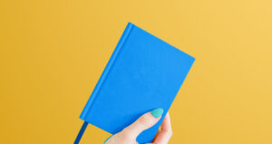Mão segurando livro azul sobre fundo amarelo