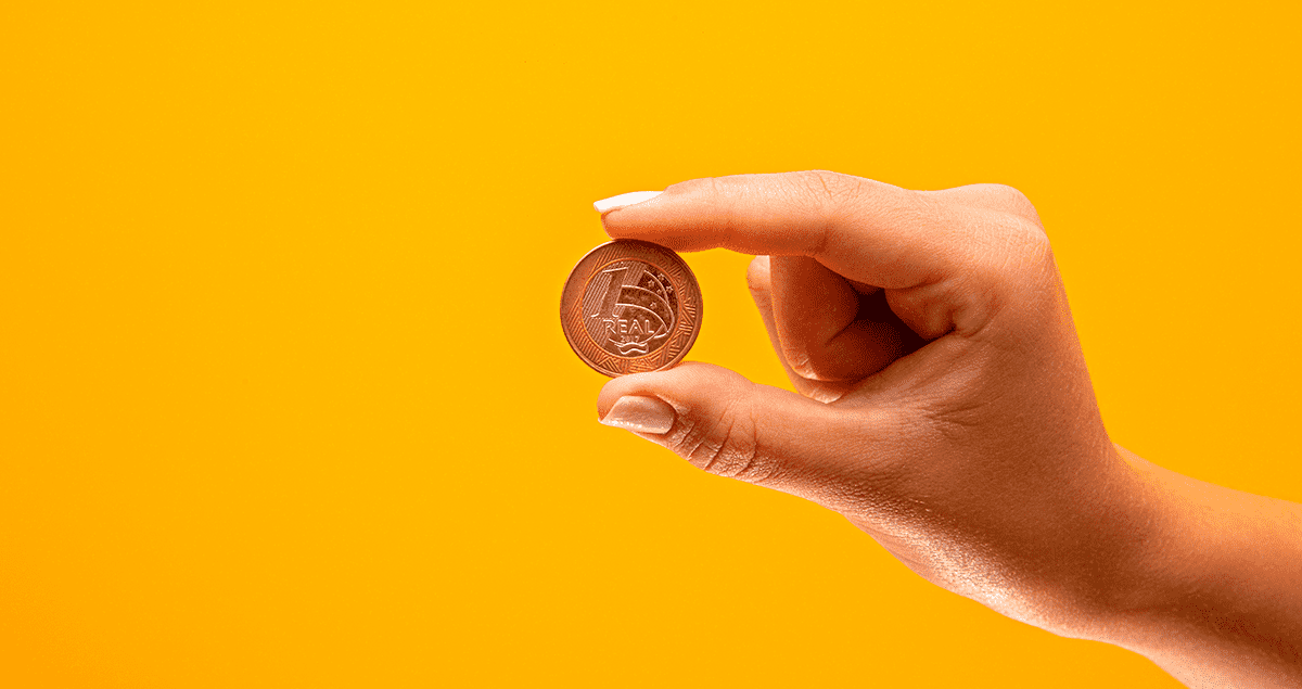 Mão segurando moeda de um real em frente a fundo amarelo