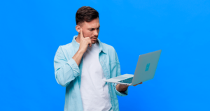 Homem em pé segurando computador nas mãos com semblante de dúvida em frente a fundo azul