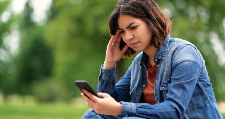 Mulher sentada ao ar livre olhando para celular com semblante preocupado