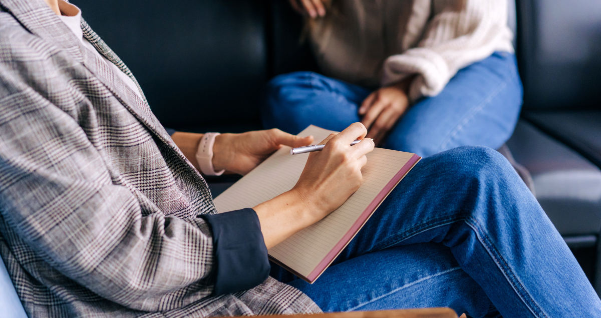 Quanto ganha um psicólogo por mês? E por consulta? 1 Pessoa sentada com caderno na mão em primeiro plano e outra pessoa sentada em sofá em segundo plano