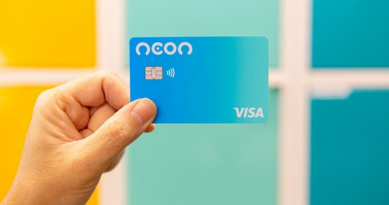 Mão segurando cartão de crédito Neon em frente a fundo colorido