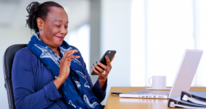 Mulher sentada em frente a computador com celular na mão e sorrindo