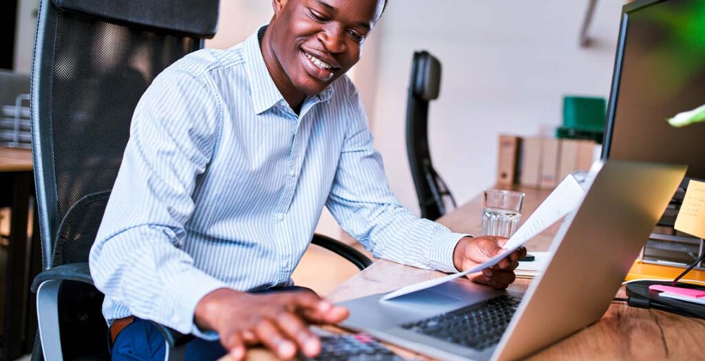 Homem segurando papel em frente a computador e digitando em calculadora