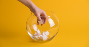 Pessoa colocando mão em pote de vidro para pegar papel em frente a fundo amarelo