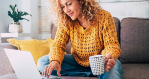 Mulher sentada em sofá e mexendo em computador com caneca na mão