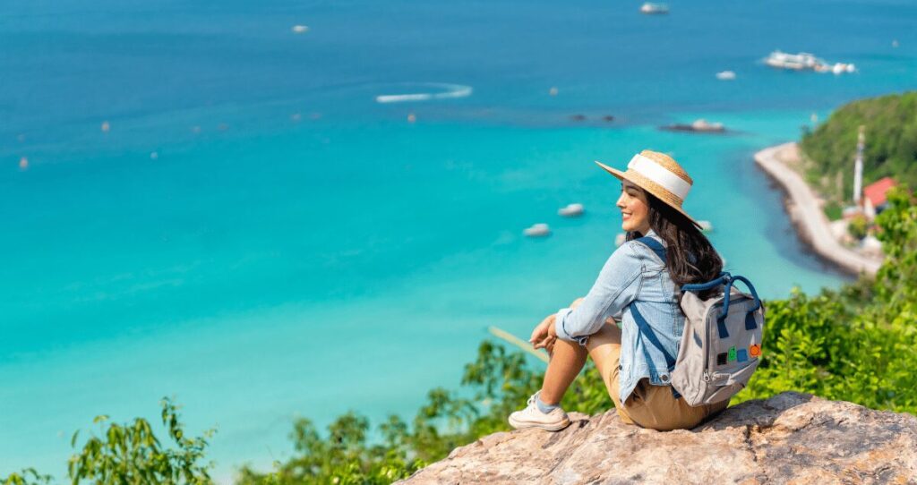 Mulher de chapéu e mochila sentada em pedra olhando para o mar