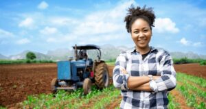 Mulher de braços cruzados sorrindo em frente a campo de agricultura com trator ao fundo