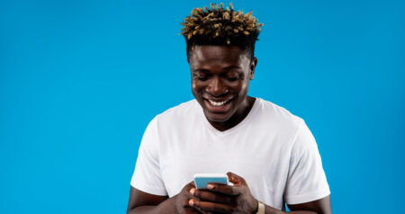 Homem mexendo em celular e sorrindo em frente a fundo azul