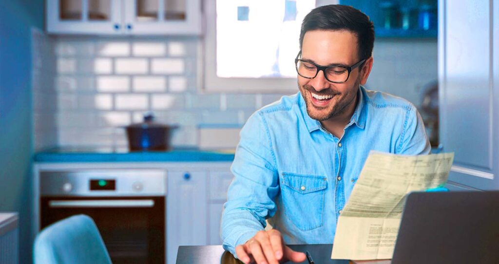 Homem segurando documento em frente a computador e sorrindo