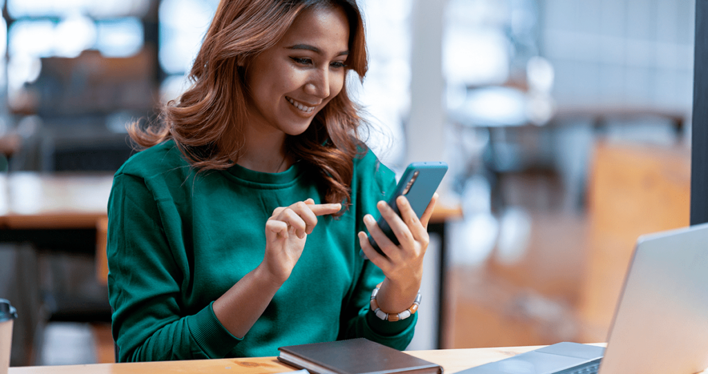 Mulher sorrindo enquanto segura celular nas mãos em frente a computador