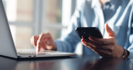 Pessoa digitando em computador com uma mão e segurando celular com a outra