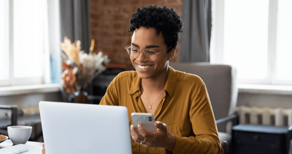 Mulher sorrindo segurando celular na mão em frente a computador