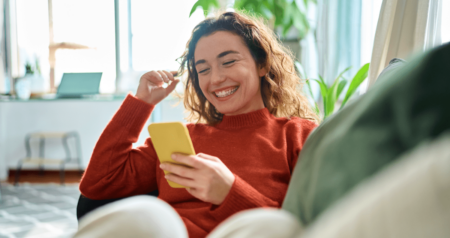 Mulher sentada em sofá segurando celular na mão e sorrindo