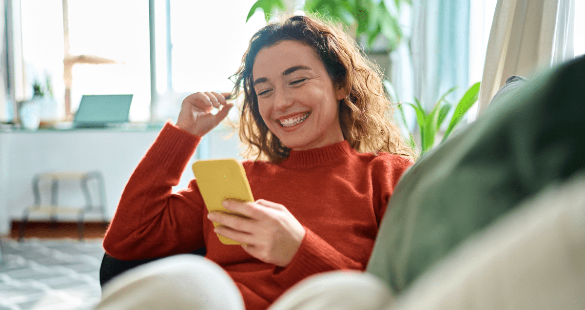 Mulher sentada em sofá segurando celular na mão e sorrindo