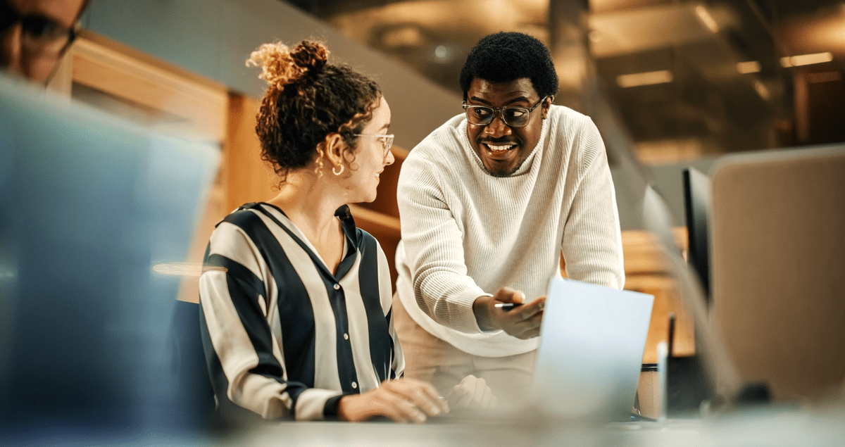 Homem em pé ao lado de mulher em escritório, ambos em frente a computador