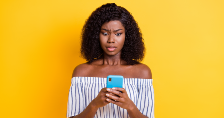 Mulher olhando para o celular com olhar de preocupação em frente a fundo amarelo